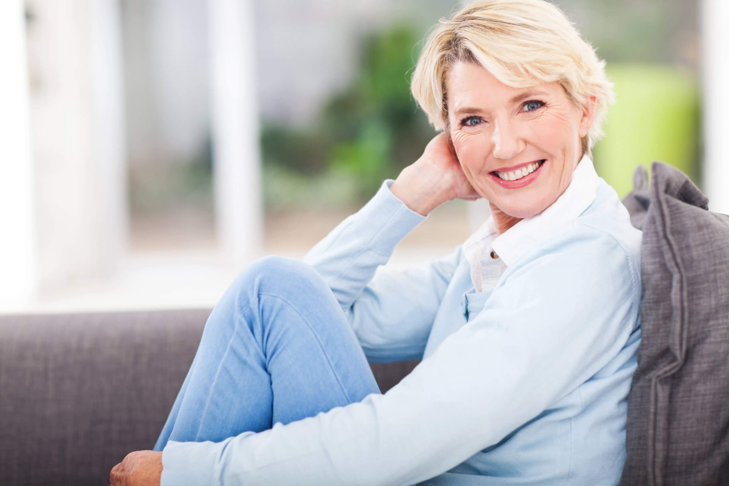 Blond Mature Female Sitting With Knee Up On Gray Couch