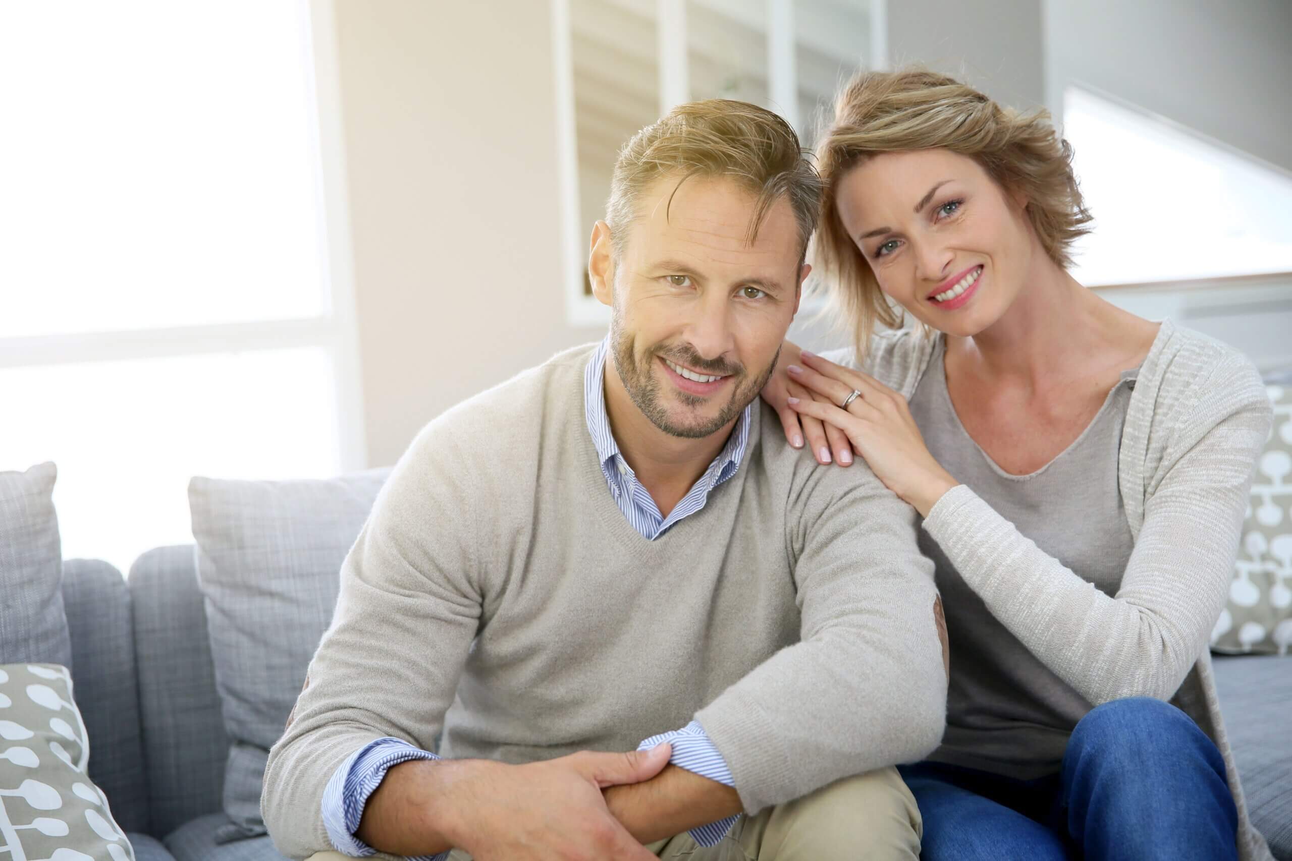 Middle Aged Couple Sitting On Gray Couch Smiling At Camera